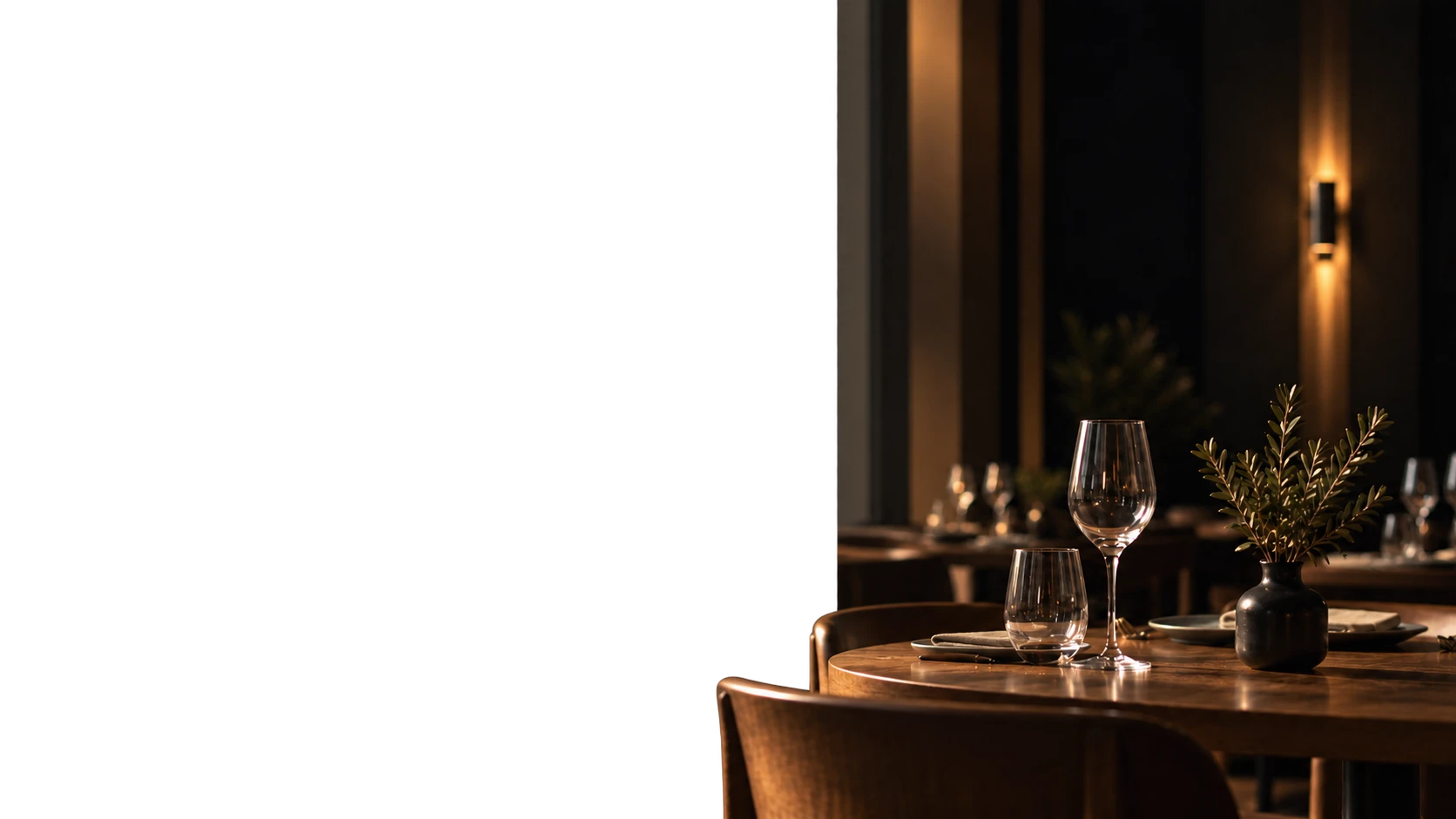 Sala de restaurante en penumbra con una mesa preparada, copas y una ramita de olivo.